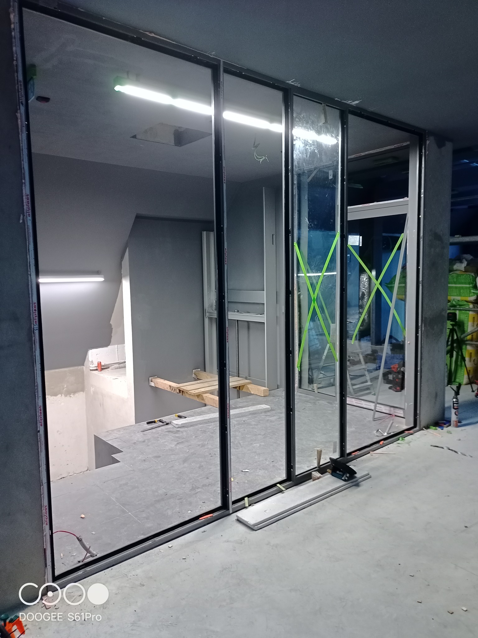 Wnętrze w trakcie prac wykończeniowych z widocznymi przesuwnymi szklanymi panelami z czarnymi ramami, zabezpieczonymi zieloną taśmą malarską, narzędzia na podłodze, surowy beton i schody w tle.