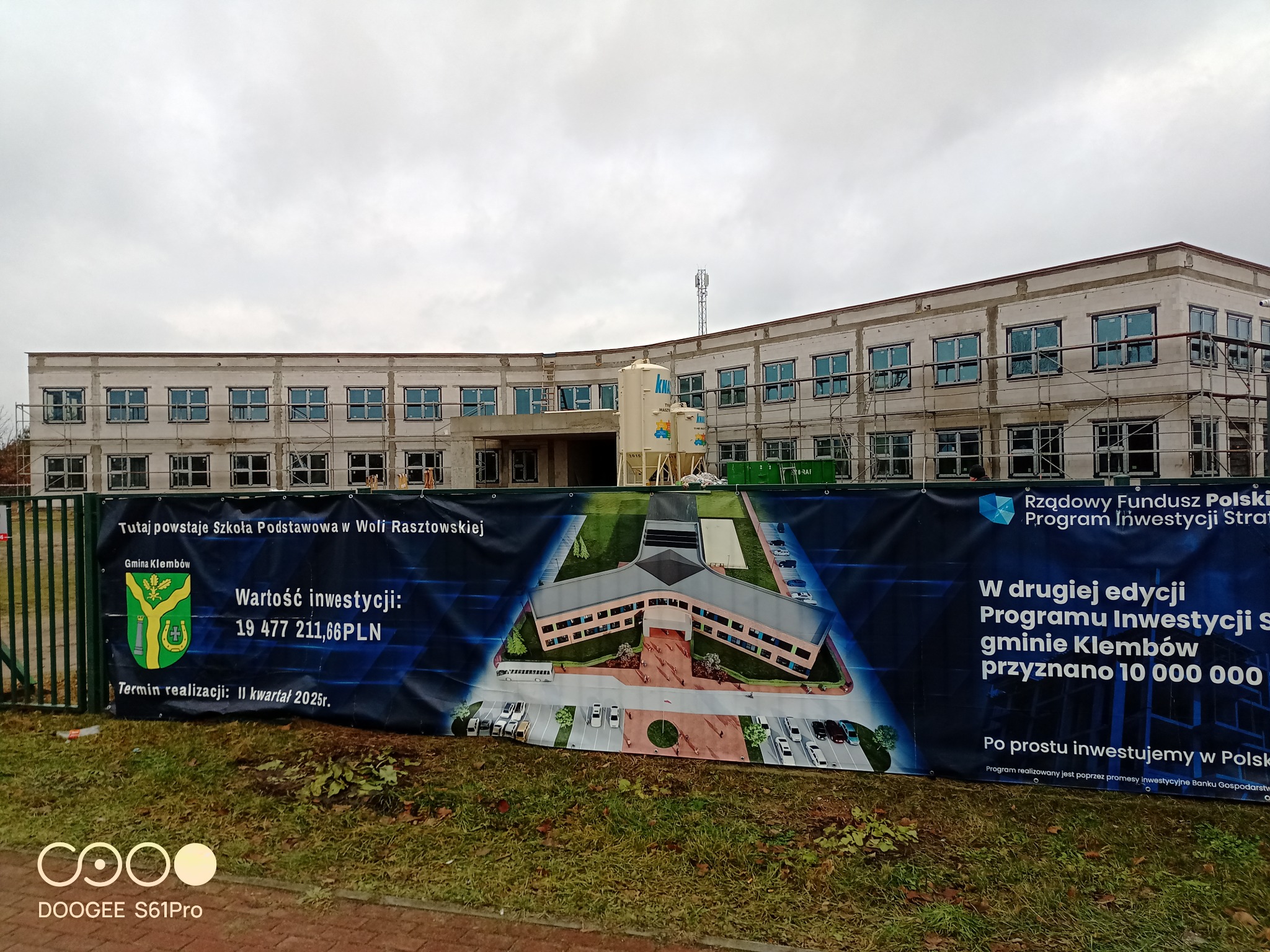 Budowa szkoły podstawowej w Woli Rasztowskiej w trakcie montażu okien, widoczny baner informacyjny z wizualizacją budynku i danymi o finansowaniu inwestycji.