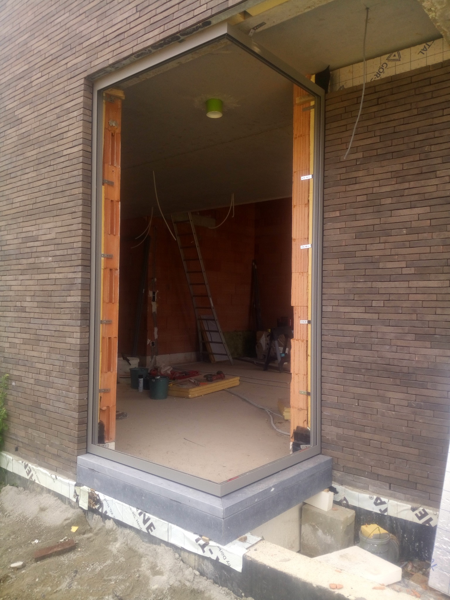 Surowy otwór okienny w ścianie z cegły klinkierowej, z widoczną konstrukcją montażową i wnętrzem w trakcie remontu, gdzie widać drabinę i narzędzia.