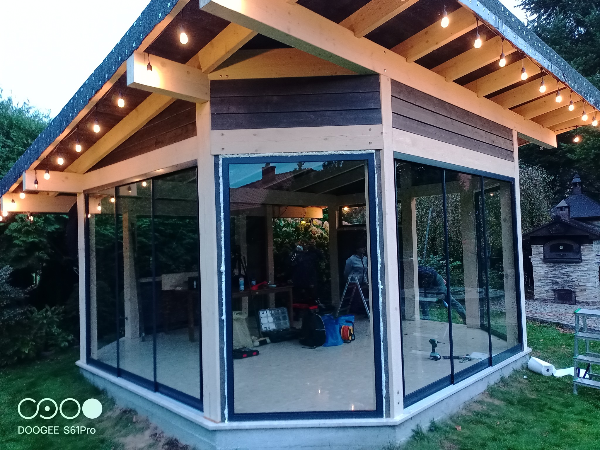 Zadaszona konstrukcja z przeszklonymi ścianami w trakcie montażu, widoczne narzędzia i osoby pracujące wewnątrz, oświetlenie w postaci wiszących żarówek, piec na zewnątrz.