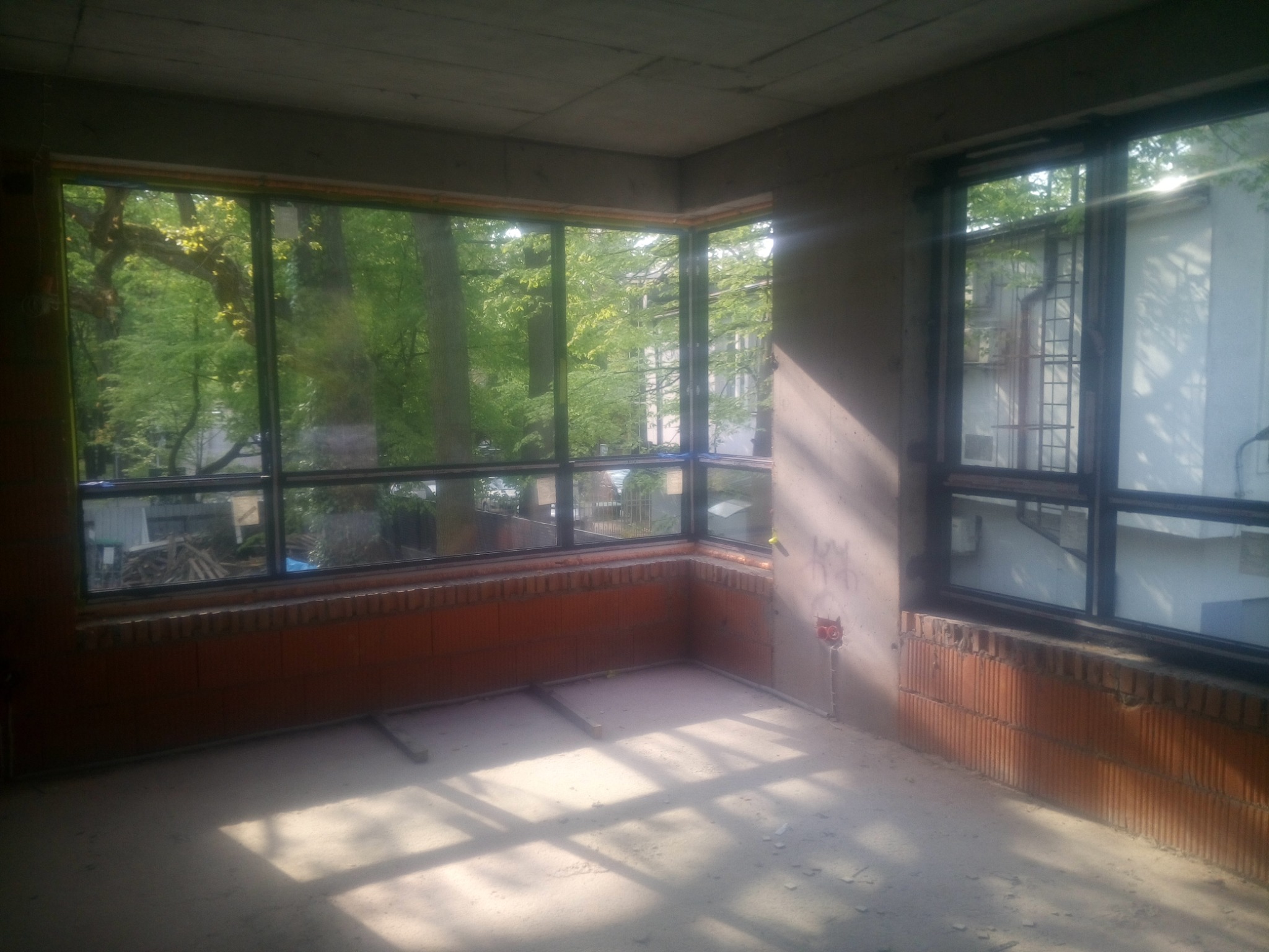 Surowy stan wnętrza z dużymi oknami z czarną ramą, widok na zieleń drzew za oknem, ściany z czerwonej cegły, wylewka betonowa na podłodze, widoczne instalacje elektryczne.