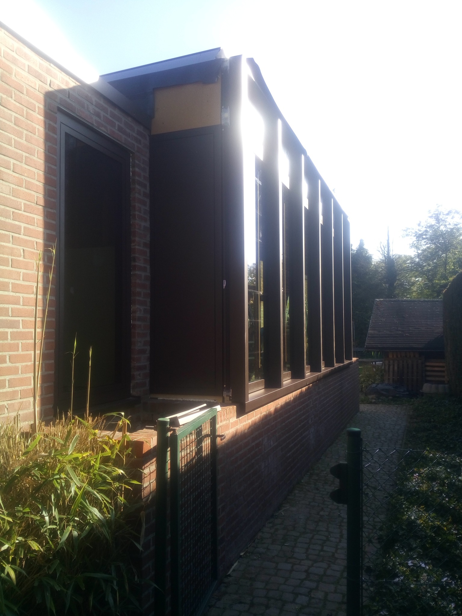 Nowoczesna bryła budynku z cegły z ciemnymi oknami i drzwiami, widok z zewnątrz, fragment ogrodu z roślinnością i ogrodzeniem.