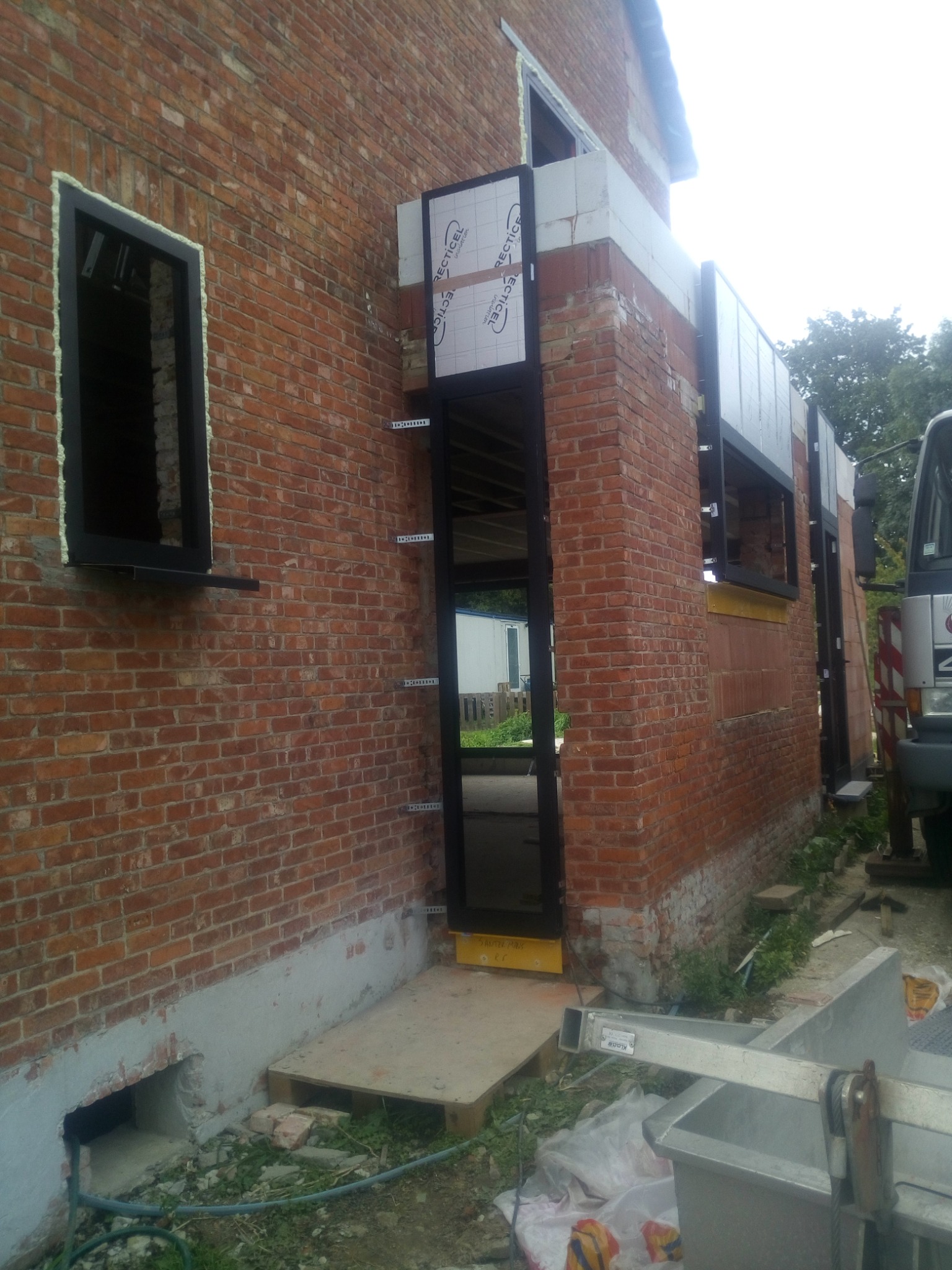 Częściowy widok murowanej ściany budynku z zamontowanymi oknami z czarnymi ramami i przygotowanymi otworami na kolejne okna, widoczne elementy rusztowania i materiały budowlane.