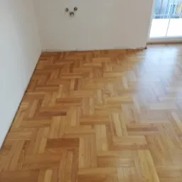 Parkiet ułożony we wzór jodełki francuskiej, widoczne ślady po listwach przypodłogowych, przygotowany do dalszych prac wykończeniowych.