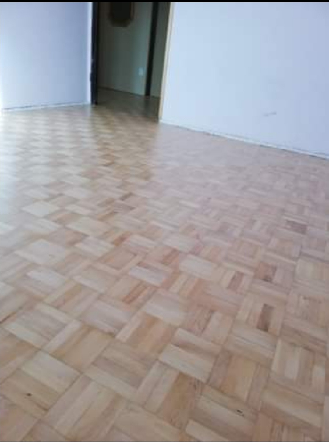 Jasny parkiet mozaikowy ułożony w jodełkę na podłodze w pustym pokoju z białymi ścianami i widocznym fragmentem progu drzwiowego do kolejnego pomieszczenia.