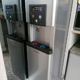 Dwa dystrybutory wody marki Ruhens, jeden srebrny, drugi grafitowy, stoją obok siebie w pomieszczeniu z kafelkową podłogą. Dystrybutory posiadają wyświetlacze i przyciski do wyboru temperatury wody.