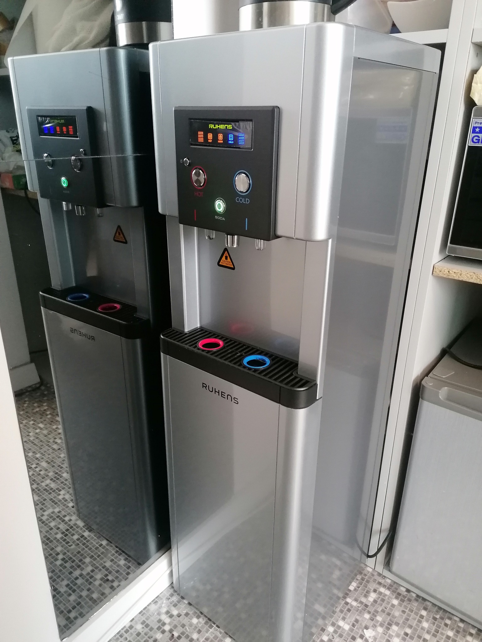 Dwa dystrybutory wody marki Ruhens, jeden srebrny, drugi grafitowy, stoją obok siebie w pomieszczeniu z kafelkową podłogą. Dystrybutory posiadają wyświetlacze i przyciski do wyboru temperatury wody.