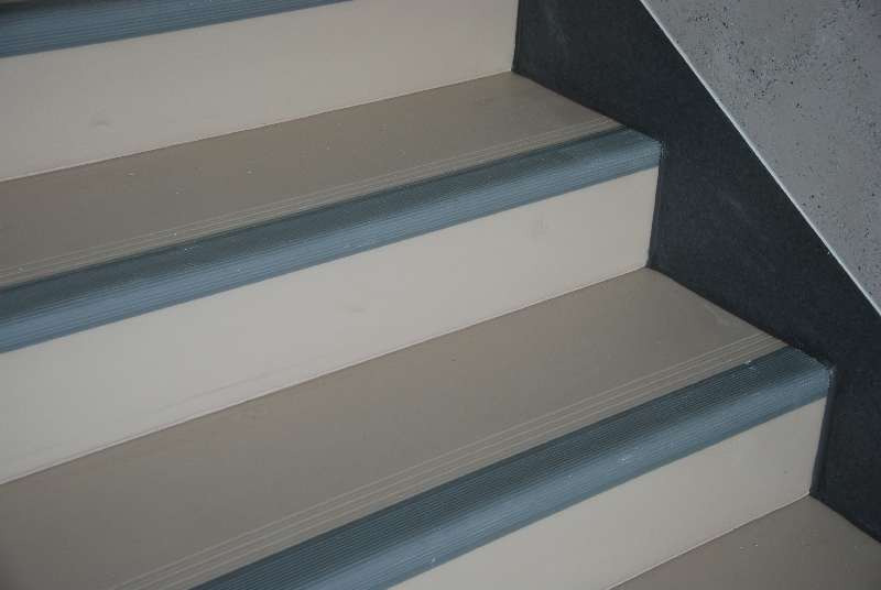 Detale wykończenia schodów z beżową wykładziną i szarymi listwami antypoślizgowymi, widoczne fragmenty ściany wykończonej w ciemnym kolorze.