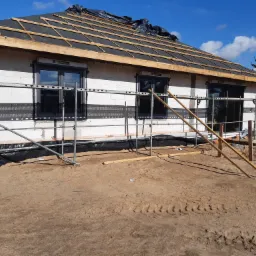 Budowa domu z białych bloczków, widoczne rusztowania, okna z czarnymi ramami, konstrukcja dachu z deskami i czarną folią, teren wokół w stanie surowym z widocznymi śladami opon.