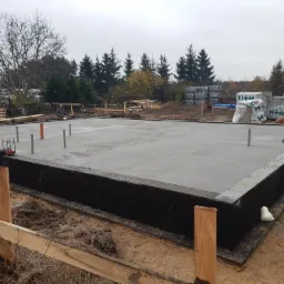 Świeżo wylana płyta fundamentowa z hydroizolacją, widoczne wystające rury i pręty, w tle zapasy bloczków H+H i pracujący robotnicy.