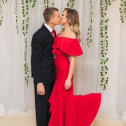 Para całująca się na tle białej zasłony z girlandami z zielonych liści i światełek; kobieta w czerwonej sukni z odkrytymi ramionami, mężczyzna w czarnym garniturze z czerwoną muchą.