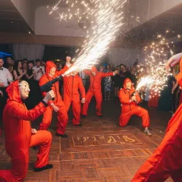 Grupa osób w czerwonych kombinezonach i maskach Dalego, imitujących napad, wystrzeliwuje iskry na parkiecie podczas imprezy, goście w tle klaszczą.