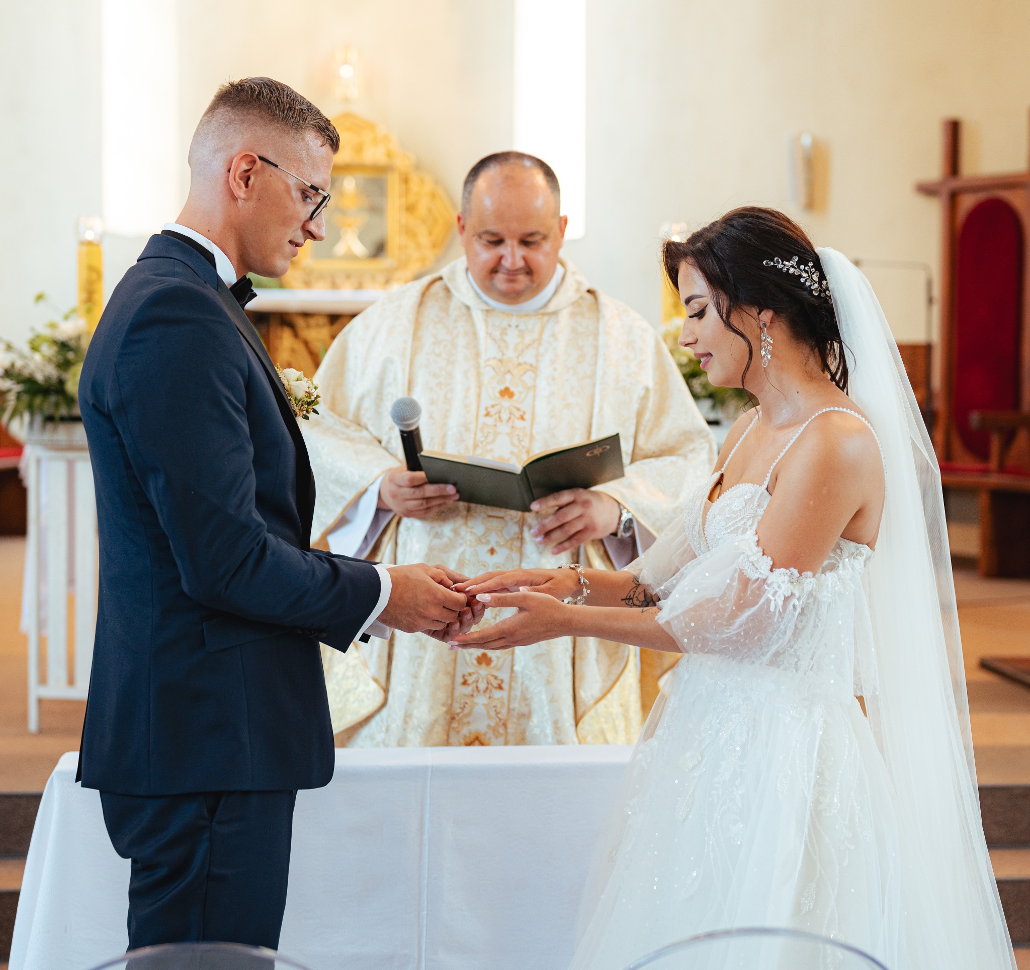 Para młoda wymienia obrączki podczas ceremonii ślubnej w kościele, w tle ksiądz z mikrofonem i księgą.