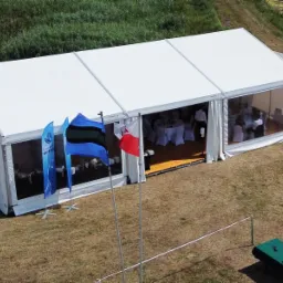 Duży, biały namiot imprezowy z przezroczystymi ścianami bocznymi, widok z góry na wnętrze z ustawionymi stołami i krzesłami, na zewnątrz powiewające flagi i przygotowane stanowisko.