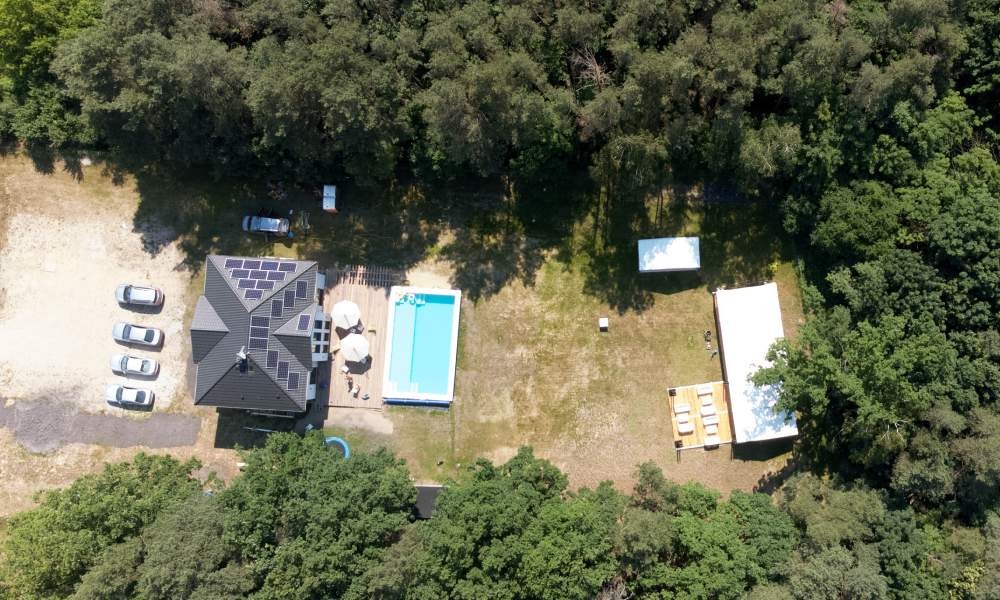 Widok z lotu ptaka na posesję z domem z panelami słonecznymi, basenem i białymi namiotami imprezowymi na trawniku otoczonym drzewami.