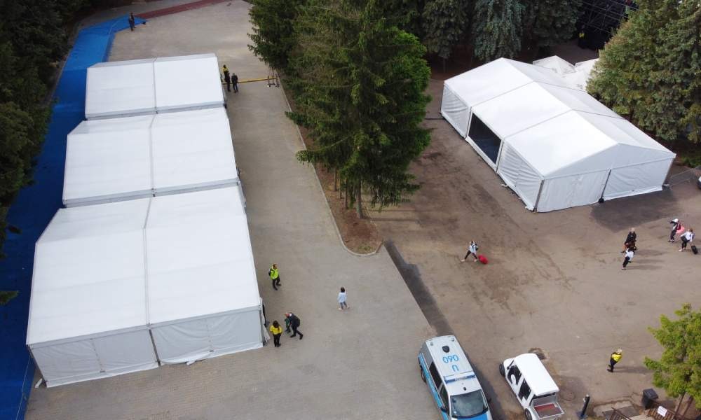 Widok z góry na rozstawione białe namioty eventowe, z ludźmi przechodzącymi obok i stojącymi samochodami na utwardzonym placu.