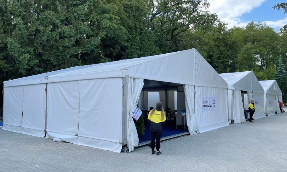 Rząd białych namiotów z wejściami, widoczne osoby i drzewa w tle, prawdopodobnie podczas wydarzenia plenerowego.