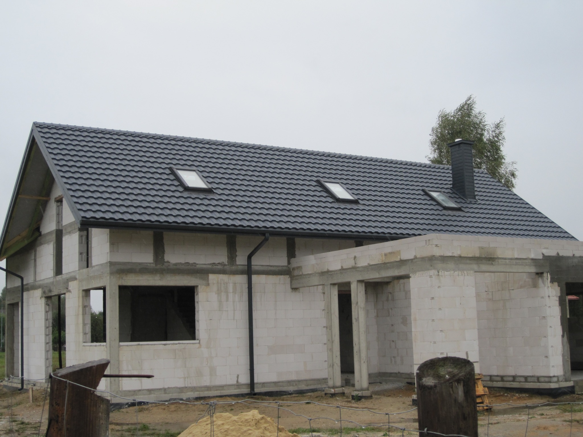Dom w trakcie budowy z czarną dachówką i trzema oknami dachowymi, widoczne pustaki i elementy konstrukcyjne.