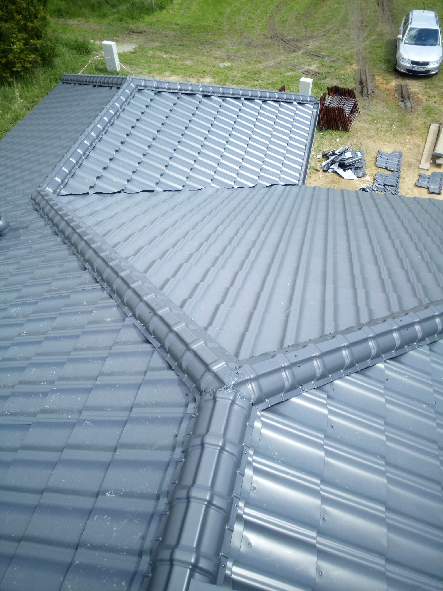 Nowo położony, szary dach z blachodachówki modułowej z widocznymi elementami wykończeniowymi, z perspektywy z góry; w tle zaparkowany srebrny samochód osobowy.