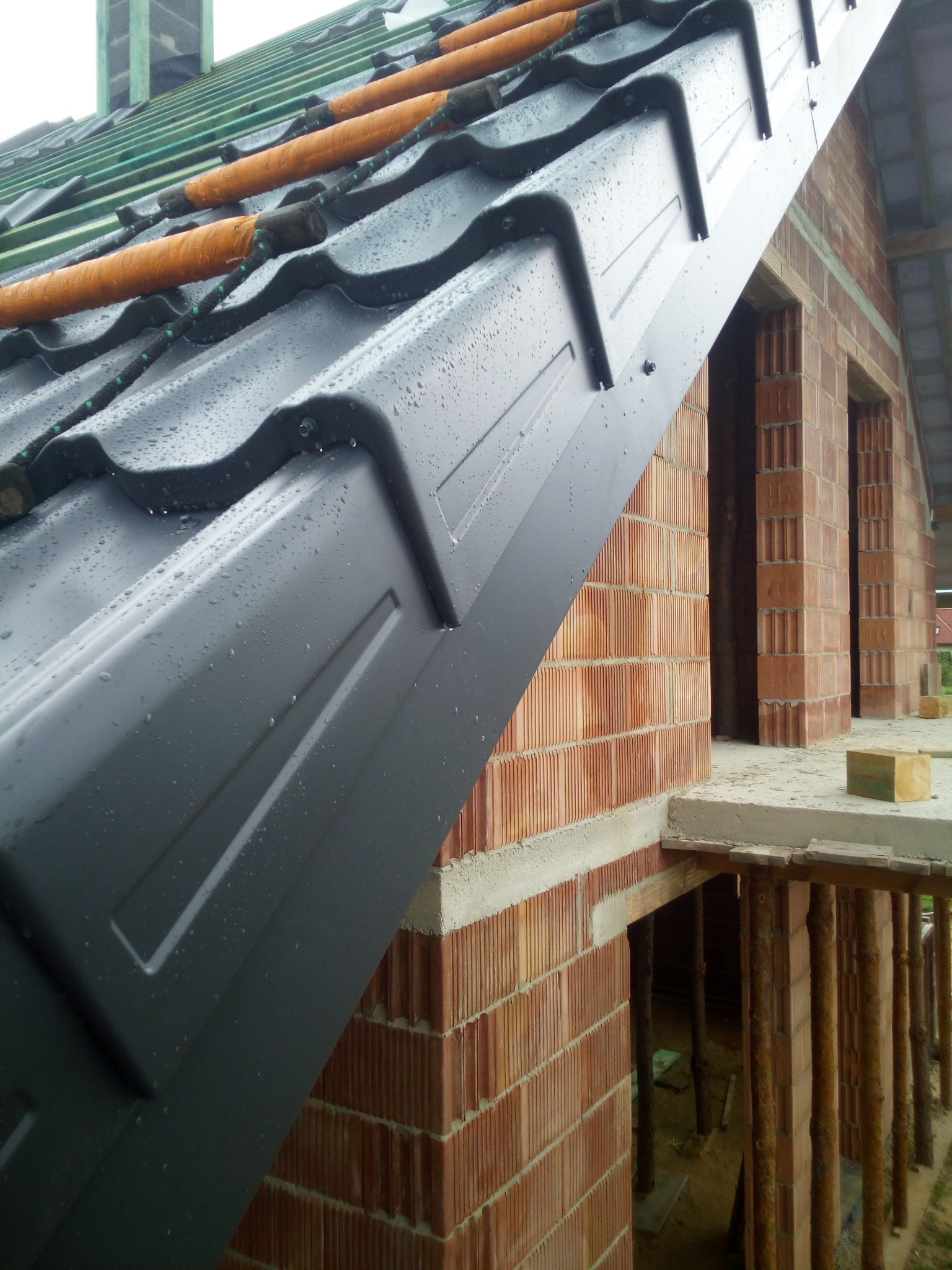 Fragment dachu pokrytego grafitową blachodachówką, na której widoczne są krople deszczu; w tle surowa ściana budynku z czerwonej cegły i elementy konstrukcji drewnianej.
