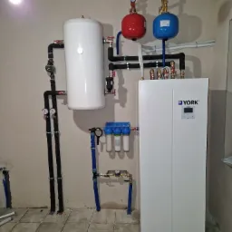 Biały zbiornik buforowy i pompa ciepła YORK z podłączeniami hydraulicznymi, manometry, filtry wody, instalacja w pomieszczeniu z płytkami podłogowymi i surowymi ścianami.