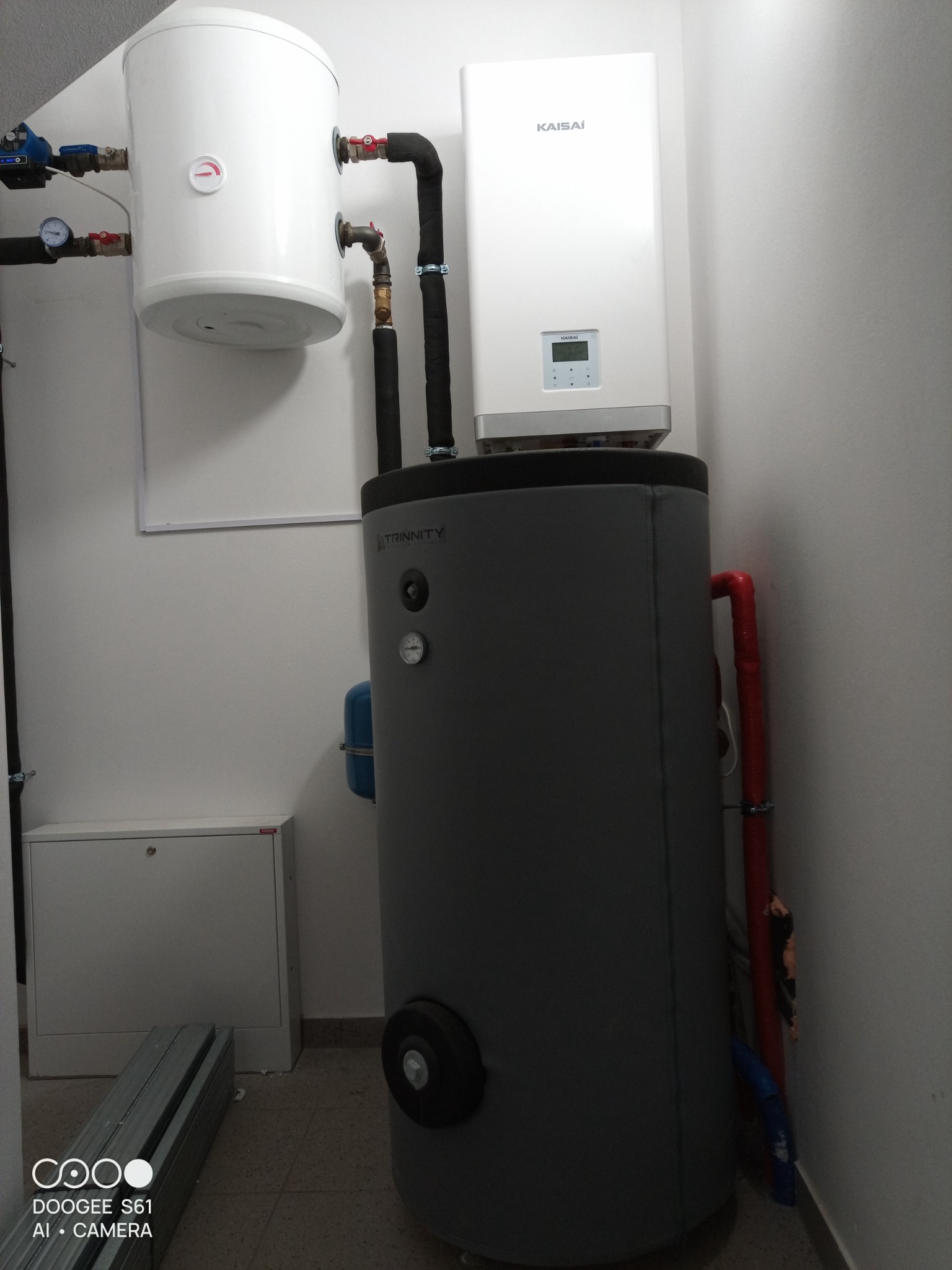 Biały zasobnik ciepłej wody, szary akumulator ciepła Trinity i biała pompa ciepła Kaisai zamontowane w kotłowni z białymi ścianami.