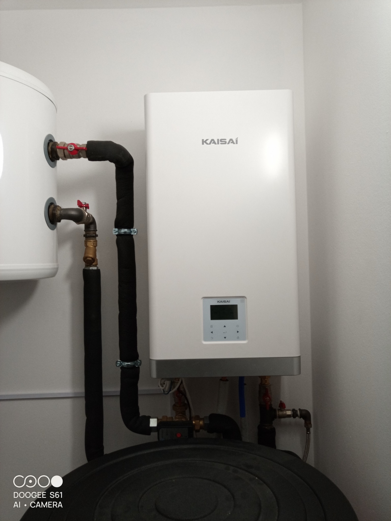 Nowoczesny, biały piec Kaisai zamontowany w kotłowni, widoczne rury w izolacji, zawory i czarny zbiornik akumulacyjny.