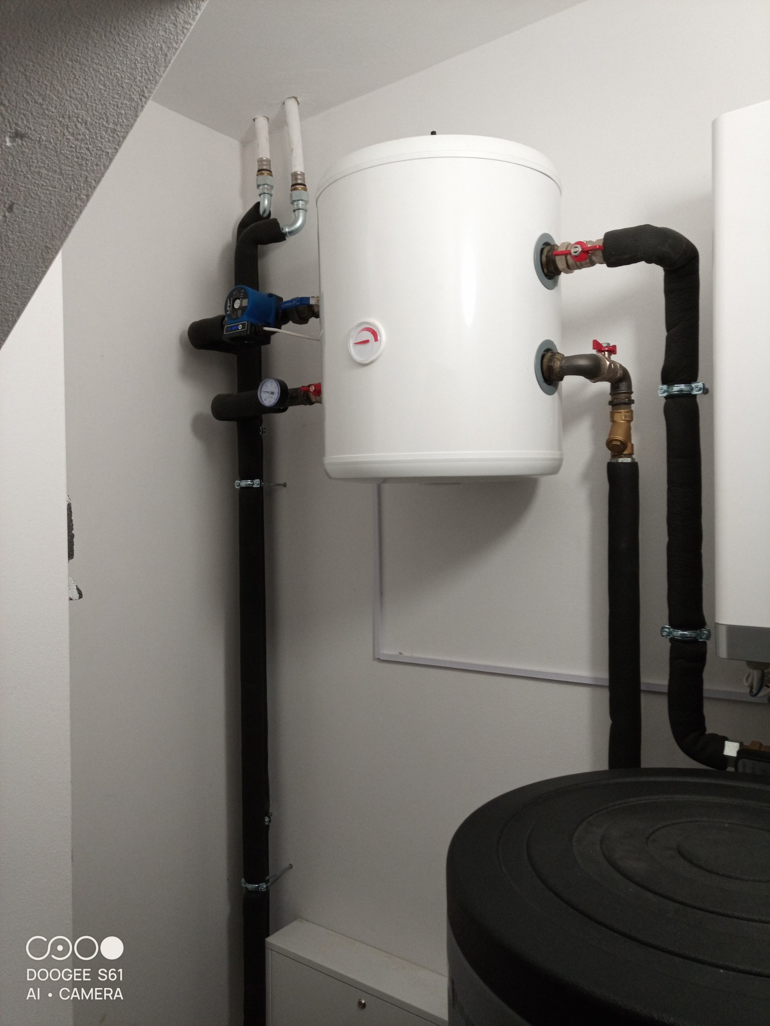 Biały zasobnik ciepłej wody użytkowej z czarną izolacją na rurach, zamontowany w pomieszczeniu technicznym z szarą ścianą.