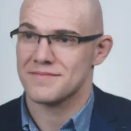 Portret łysiejącego mężczyzny w okularach i niebieskiej marynarce na jasnym tle.