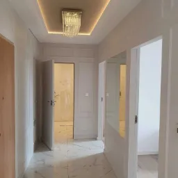 Hallway with white marble-patterned floor tiles, wainscoting, and a crystal chandelier under a recessed, illuminated ceiling panel.