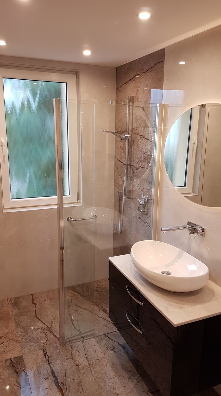 Nowoczesna łazienka z kabiną prysznicową ze szklanymi drzwiami z chromowanym uchwytem, umywalką nablatową o owalnym kształcie na czarnej szafce z marmurowym blatem, okrągłym lustrem...