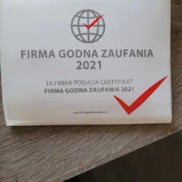 Certyfikat 'Firma Godna Zaufania 2021' z symbolem globu i czerwonym znakiem akceptacji, leżący na drewnianej powierzchni.