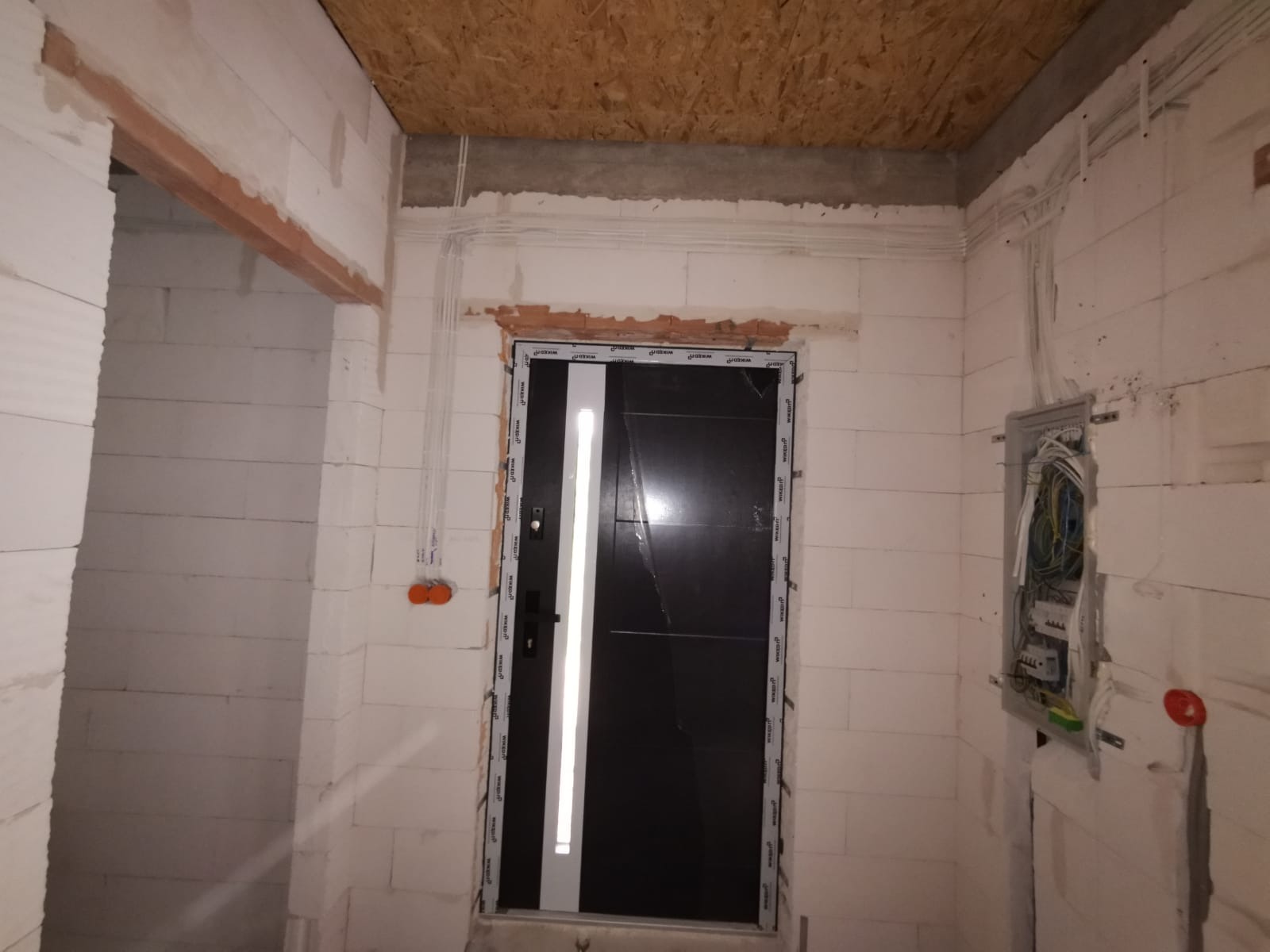 Wnętrze w trakcie prac wykończeniowych: widoczne białe ściany z betonu komórkowego, instalacja elektryczna w peszlach, otwarta skrzynka rozdzielcza z przewodami, oraz zamontowane, ale jeszcze...