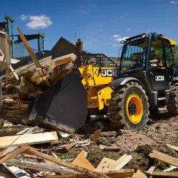 Żółta ładowarka teleskopowa JCB z czarną łyżką pełną gruzu i resztek materiałów budowlanych na placu budowy, w tle częściowo zburzony budynek.
