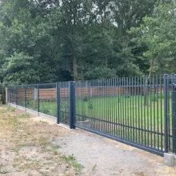 Nowoczesne, szare ogrodzenie panelowe z bramą przesuwną na podmurówce betonowej, widok z zewnątrz na tle drzew.