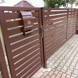 Brązowa, przesuwna brama wjazdowa z poziomymi panelami i skrzynką pocztową z numerem 10 przy ulicy Naddawki, widok z zewnątrz.