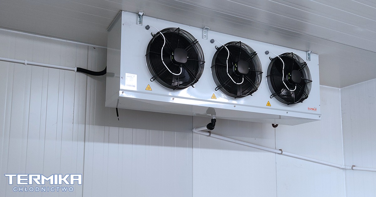 Biała jednostka chłodnicza z trzema wentylatorami zamontowana pod sufitem w chłodni, widoczne przewody i rury instalacji.