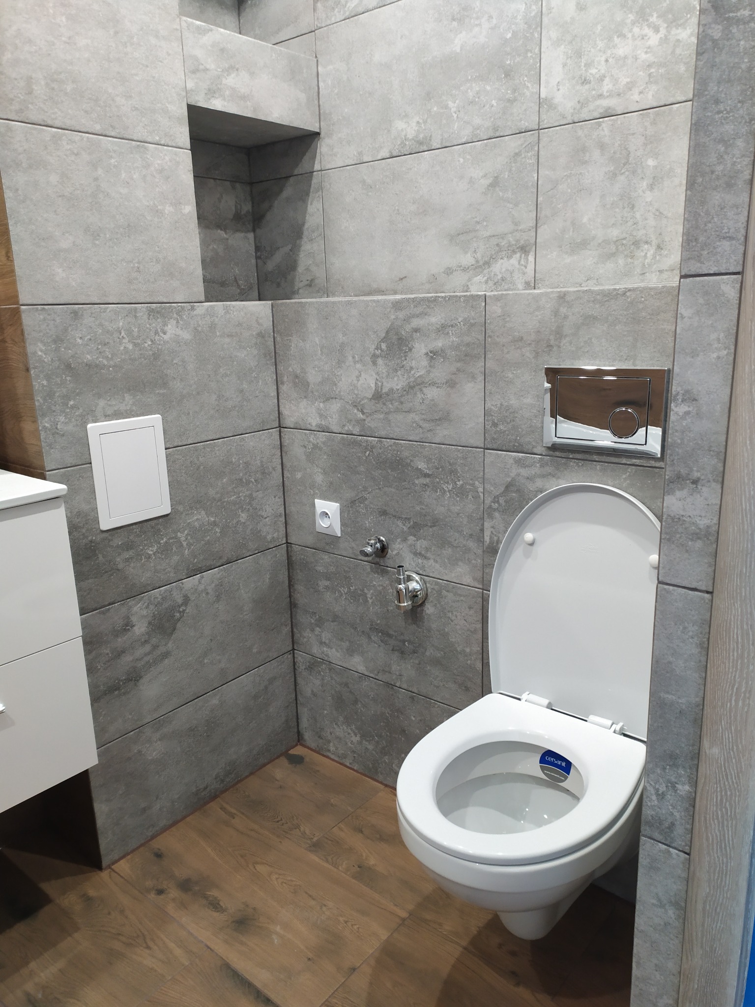 Nowoczesna toaleta wisząca z szarą, kamienną okładziną ścienną i drewnopodobną podłogą, widoczna wnęka w ścianie i biała szafka umywalkowa.