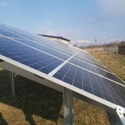 Rząd paneli słonecznych na metalowej konstrukcji, z budynkiem i drzewami w tle pod błękitnym niebem.