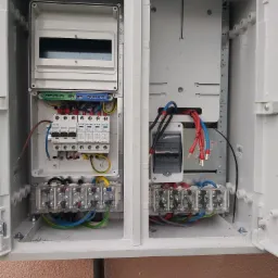 Otwarta skrzynka elektryczna z zainstalowanymi zabezpieczeniami i listwami zaciskowymi, widoczne kable w kolorach niebieskim, czerwonym, czarnym i zielono-ż&oacute;łtym.