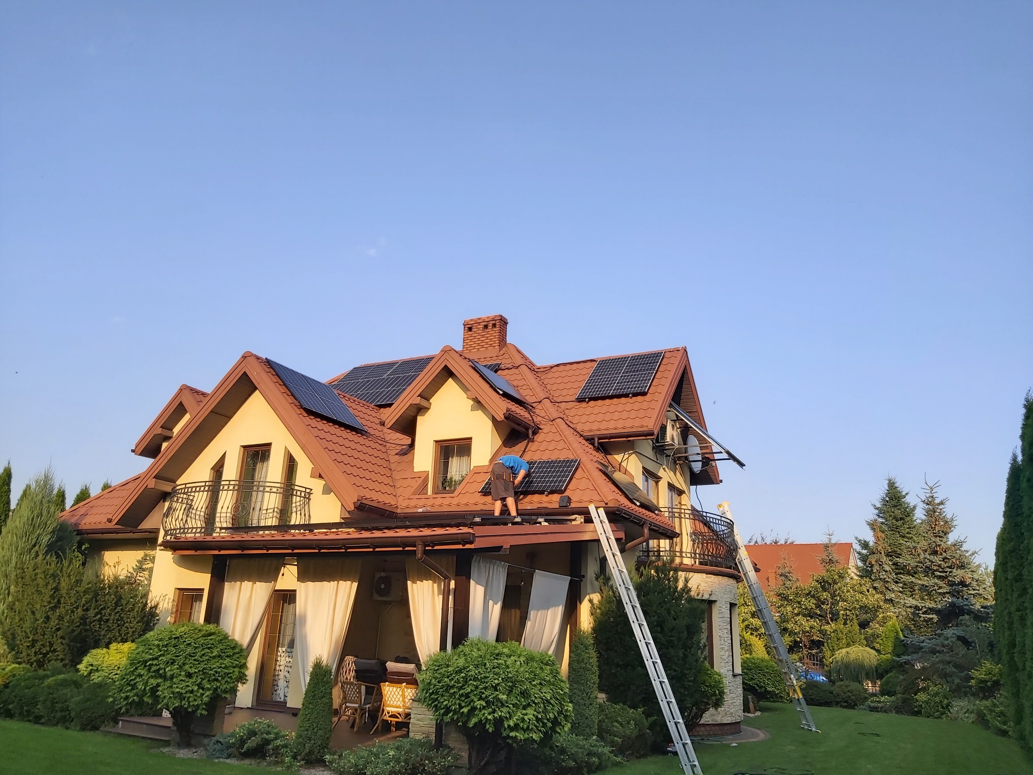 Montaż paneli fotowoltaicznych na dachu dwupiętrowego domu z czerwoną dachówką, widoczne dwie drabiny opierające się o budynek, instalator w niebieskiej koszulce pochylony nad panelami.