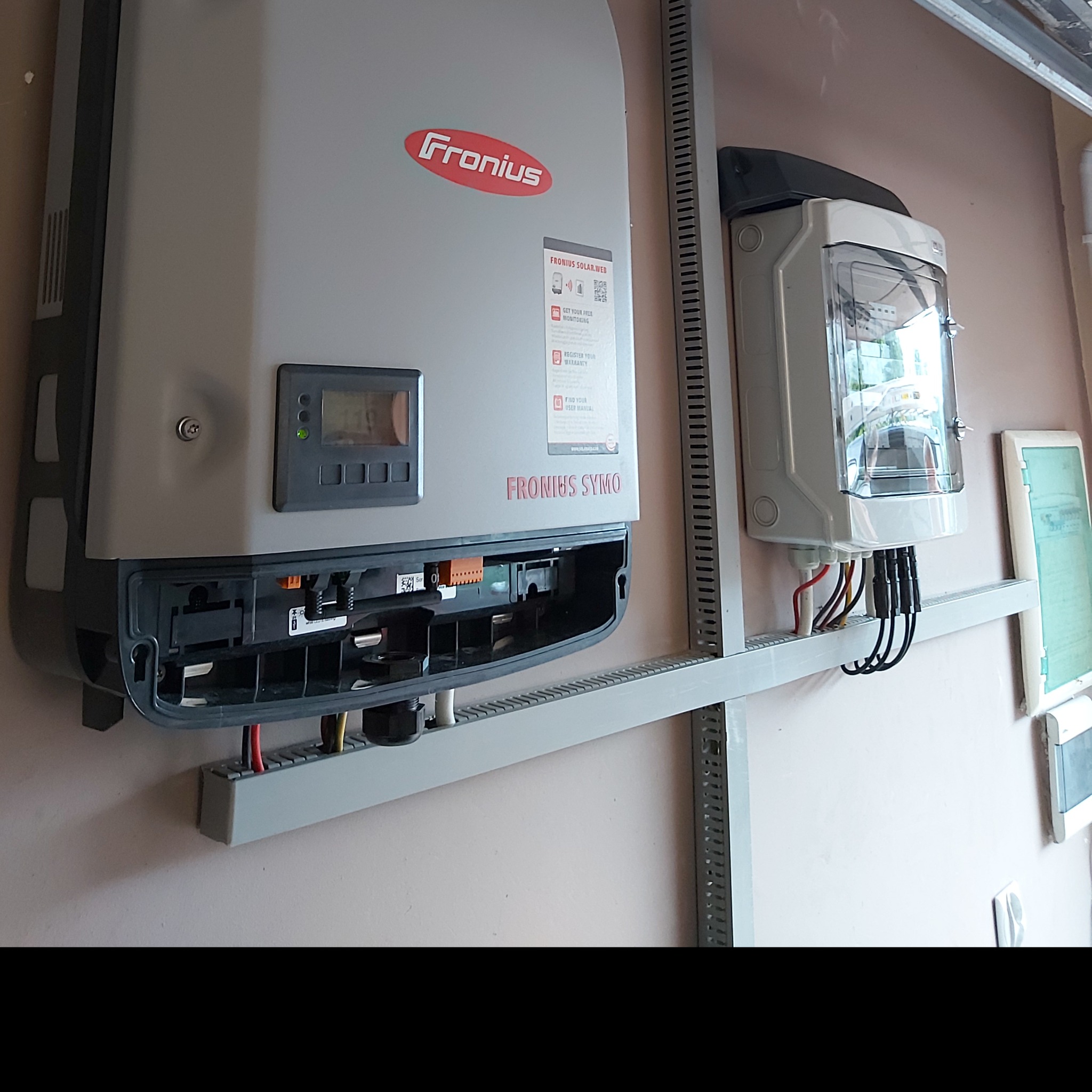 Szary falownik Fronius Symo zamontowany na metalowej konstrukcji obok skrzynki elektrycznej z widocznymi przewodami, element instalacji fotowoltaicznej.