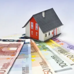 Model domu z czerwonymi ścianami i szarym dachem, umieszczony na wachlarzu banknotów euro o różnych nominałach, na białym tle.