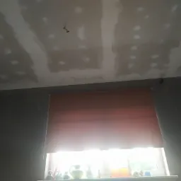 Przyciemnione wnętrze pokoju z widocznym fragmentem sufitu pokrytego płytami kartonowo-gipsowymi w trakcie szpachlowania, z widocznymi łączeniami i punktowymi poprawkami, oraz fragment ściany...