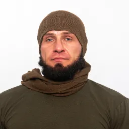 Portret mężczyzny z brodą, ubranego w zieloną koszulkę, brązową czapkę i szalik na białym tle.