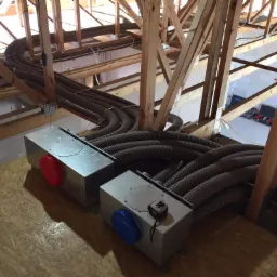 Instalacja wentylacyjna z dwoma metalowymi skrzynkami z czerwonym i niebieskim zakończeniem, liczne rury wentylacyjne w przestrzeni poddasza z drewnianą konstrukcją więźby dachowej.