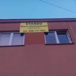 Żółty szyld z napisem 'STANDO Biuro Rachunkowe Adam Kopera tel. 668 717 004' umieszczony na brązowym budynku z dwoma oknami, widoczne przewody elektryczne na tle błękitnego nieba.