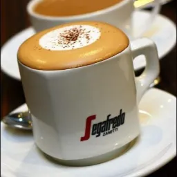 Filiżanka cappuccino marki Segafredo z posypką kakaową na białym spodku, w tle druga filiżanka z kawą latte.