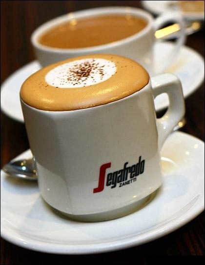 Filiżanka cappuccino marki Segafredo z posypką kakaową na białym spodku, w tle druga filiżanka z kawą latte.