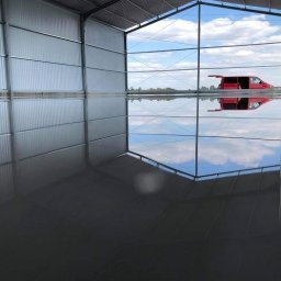 JUR-BUD Jakub Juraszek - Błyszcząca, szara posadzka wewnątrz metalowej hali z widokiem na niebo i czerwoną furgonetkę przez szklaną ścianę.